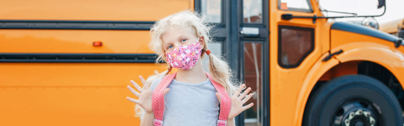 Caucasian girl in face mask standing by yellow school bus. new normal. web banner header.