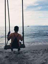 Rear view of man sitting on swing at beach against sky