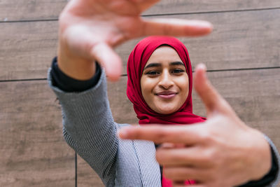 Low angle of cheerful arab female in hijab looking at camera and making frame gesture with hands while standing in street