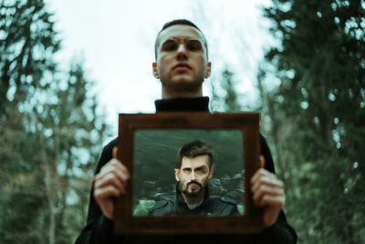 Portrait of young man holding mirror with reflection of male against trees