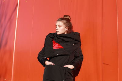 Fashionable teenage girl wearing black warm clothing standing against red wall during sunny day