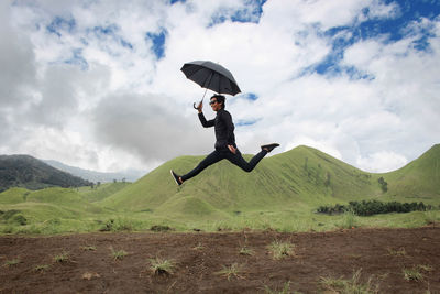 Man holding umbrella while jumping on field against mountain