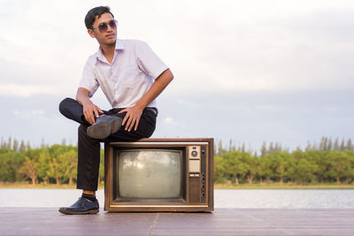 Teenage boy sitting on television set at lakeshore against sky
