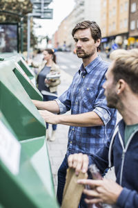 Young man with friends putting recyclable materials into recycling bins