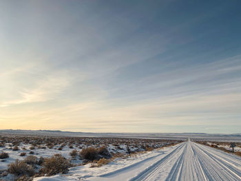 Snow covered road amidst field against sky during winter