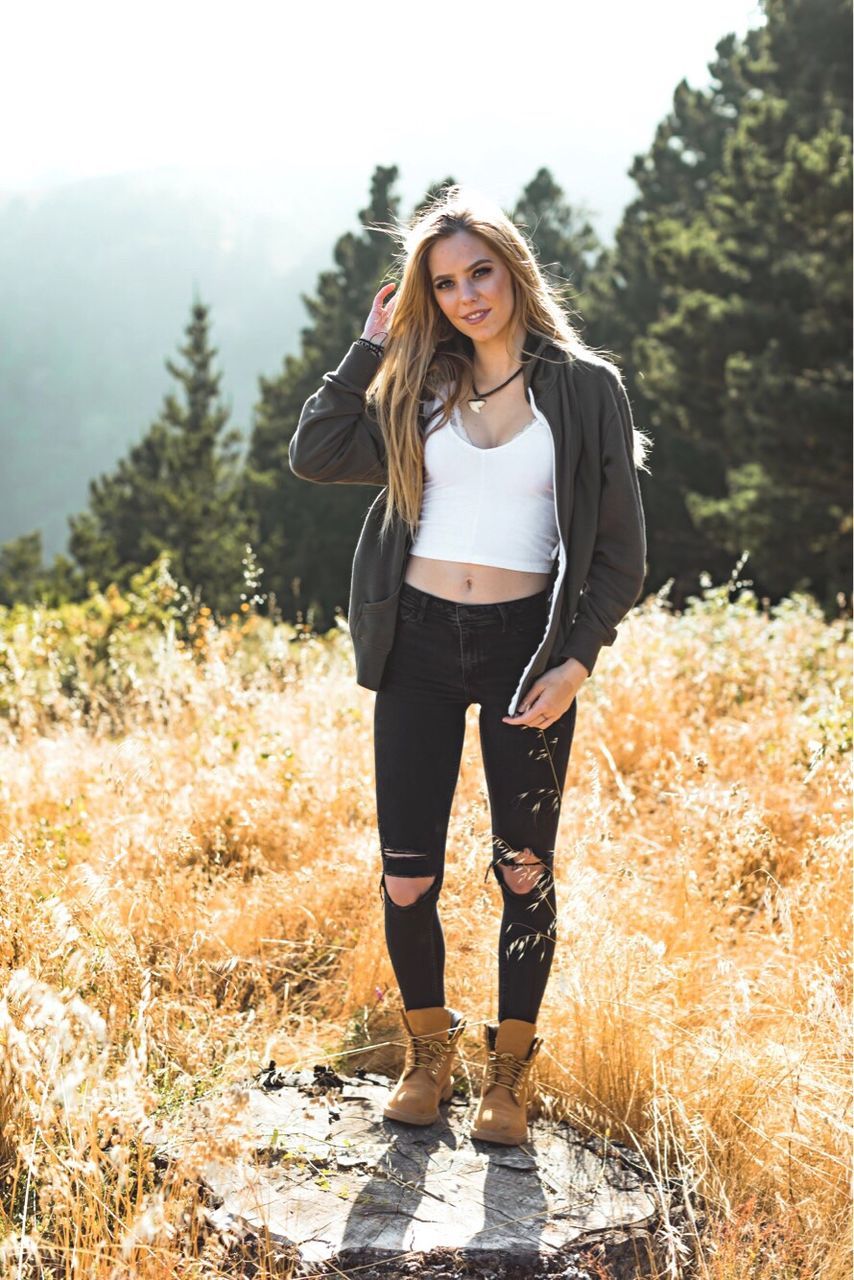 tree, one person, real people, standing, young women, front view, young adult, nature, casual clothing, leisure activity, lifestyles, forest, beautiful woman, full length, day, looking at camera, outdoors, field, hands in pockets, growth, plant, portrait, smiling, beauty in nature, sky