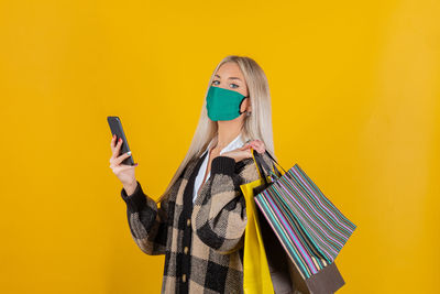 Young woman wearing mask using mobile phone against yellow background