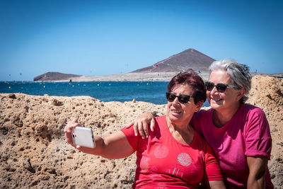 Friends taking selfie with smart phone while sitting against sea