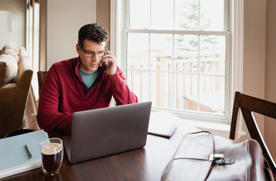 Man on cellphone working from home using a computer at a dining table.