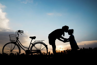 Silhouette woman kissing daughter by bicycle on field against sky during sunset