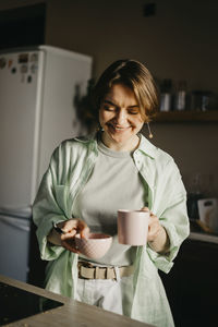 Smiling woman holding coffee cup and bowl in kitchen at home