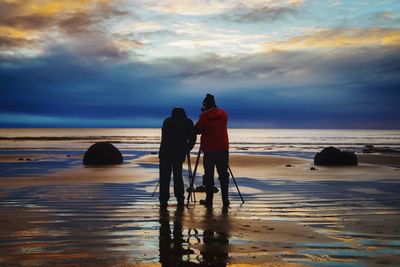 Rear view of photographers photographing on shore at beach during sunset