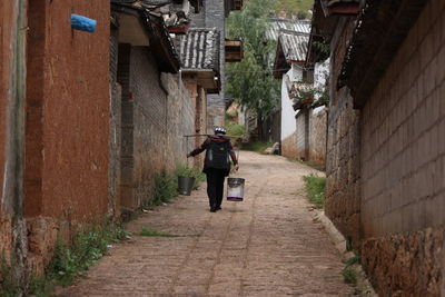Rear view of man walking on footpath carrying buckets amidst houses