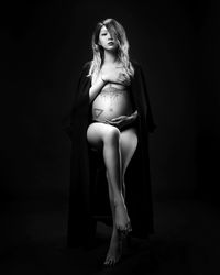 Portrait of shirtless pregnant woman sitting against black background