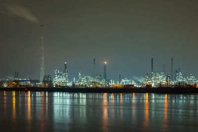 Illuminated factory by buildings against sky at night