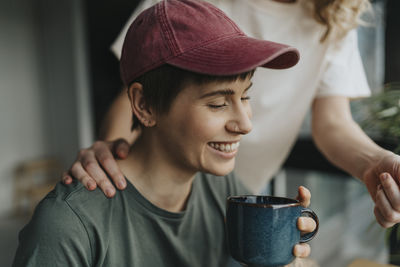 Happy lesbian woman wearing cap holding coffee cup with girlfriend's hand on shoulder