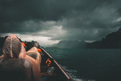 Rear view of people in boat on sea against cloudy sky