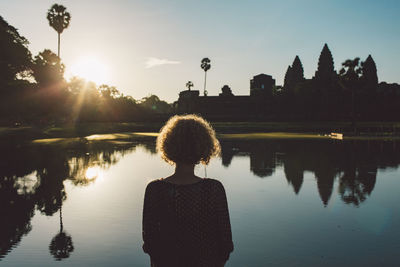 Rear view of mid adult woman standing by lake against sky during sunset