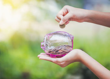 Close-up of person holding coin over transparent piggy bank 