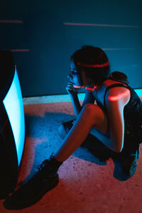 Side view of unrecognizable female model in black dress sitting on floor near glowing old television in dark studio
