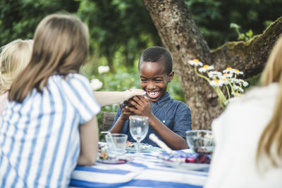 Smiling boy looking at phone while sitting with friends at dining table in backyard