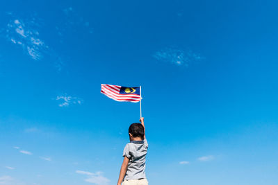 Low angle view of boy holding flag while standing against blue sky