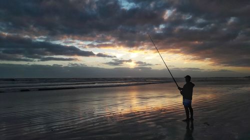 Man fishing at beach against sky during sunset