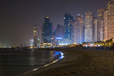 Illuminated modern buildings by sea against sky at night