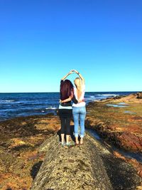 Rear view of women with arms around making heart shape over head while standing on rock at beach against sky