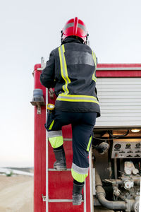 Back view of anonymous firefighter wearing red protective helmet and uniform standing on fire truck ladder and looking into the distance in daytime