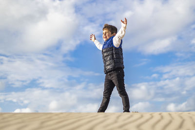 Low angle view of cute boy with arms outstretched standing on sand against sky