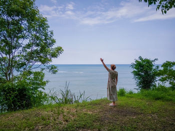 Rear view of woman with arm raised standing by sea against sky