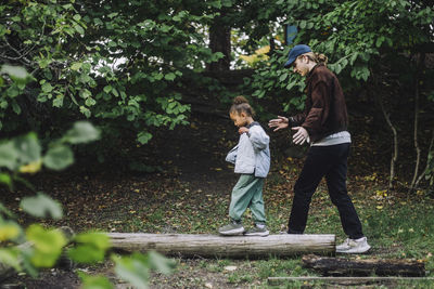 Side view of girl balancing tree on log while male teacher guiding her in park