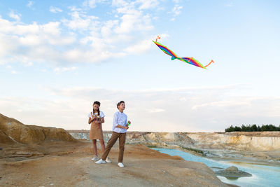 Brother and sister launch a bright big kite into the sky near a flowing river.