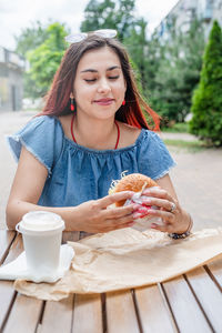 Hungry stylish woman, enjoying eating a burger outdoors, dressed in jeans shirt, wearing sunglasses