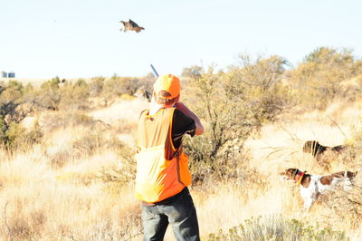 Hunter aiming pheasant flying over grassy field against sky