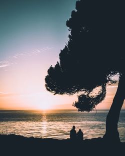 Silhouette couple sitting at beach against sky during sunset
