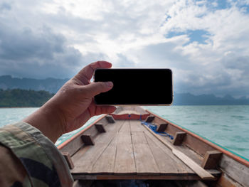 Cropped hand holding smart phone on boat in sea against sky