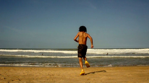 Rear view of shirtless boy running towards sea at beach against clear sky