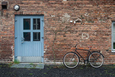 A black bike has been parked by a brick wall and a blue door. a young tree grows by the door.