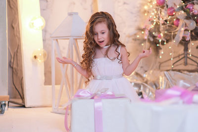 A beatiful girl with curly hair in white dress is surprising of christmas box gift near christmas 