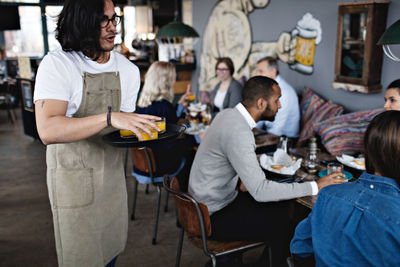 Young waiter serving drinks while standing against customers at restaurant