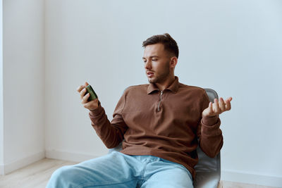 Young man using mobile phone while sitting against wall