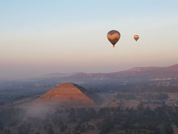 Hot air balloons flying over ancient pyramid in mist against sky during sunrise