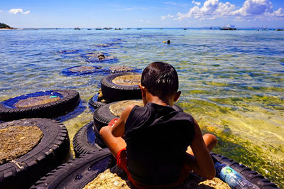 Rear view of boy sitting at beach by tiers against blue sky