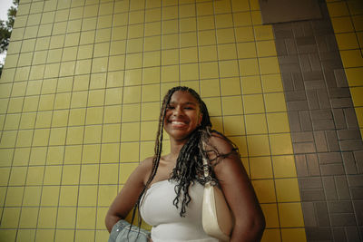 Low angle view of happy teenage girl with braided hair standing against yellow wall
