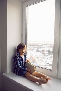 Boy child sits on the window of a house with a book in winter on a high floor