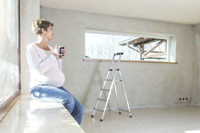 Pregnant woman holding cup while sitting by window at home