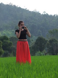 Portrait of woman photographing through mobile phone while standing on grassy field