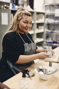 Portrait of smiling young woman with work tool and molded clay sitting at table in art class